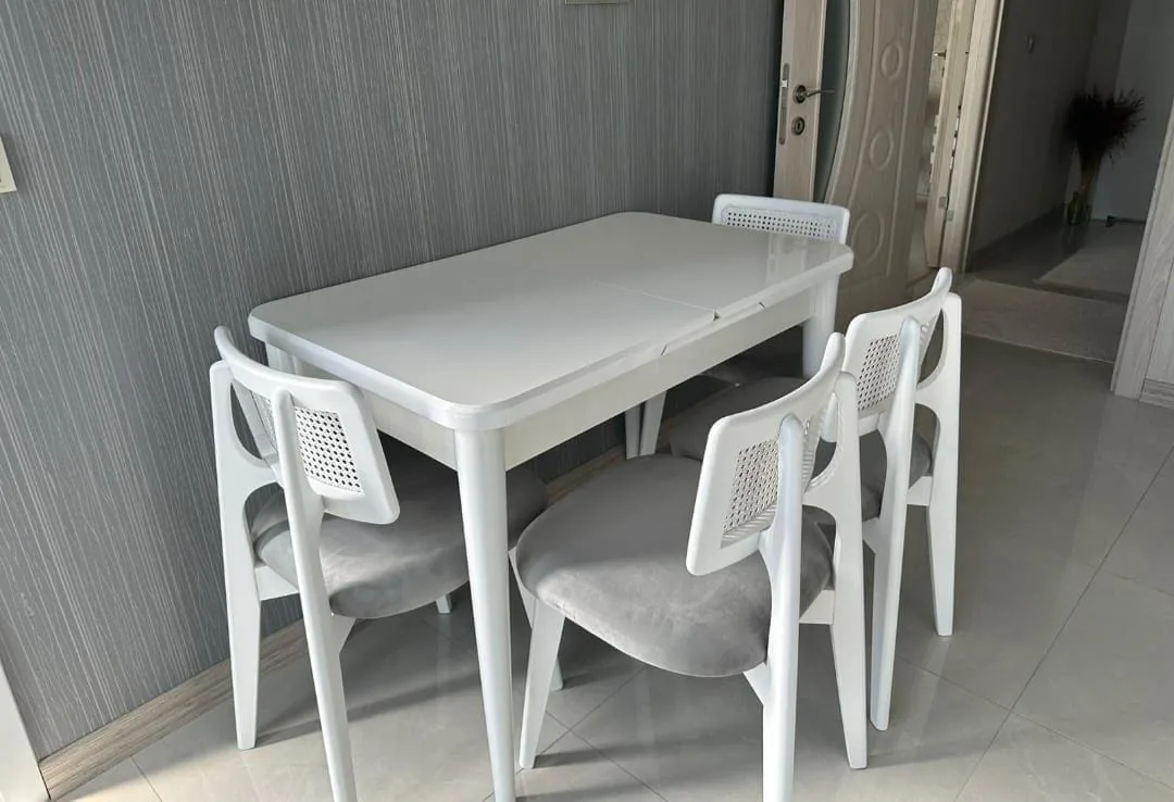 Beyaz açılır yemek masası ve dört beyaz hasır sırtlı, gri döşemeli sandalye